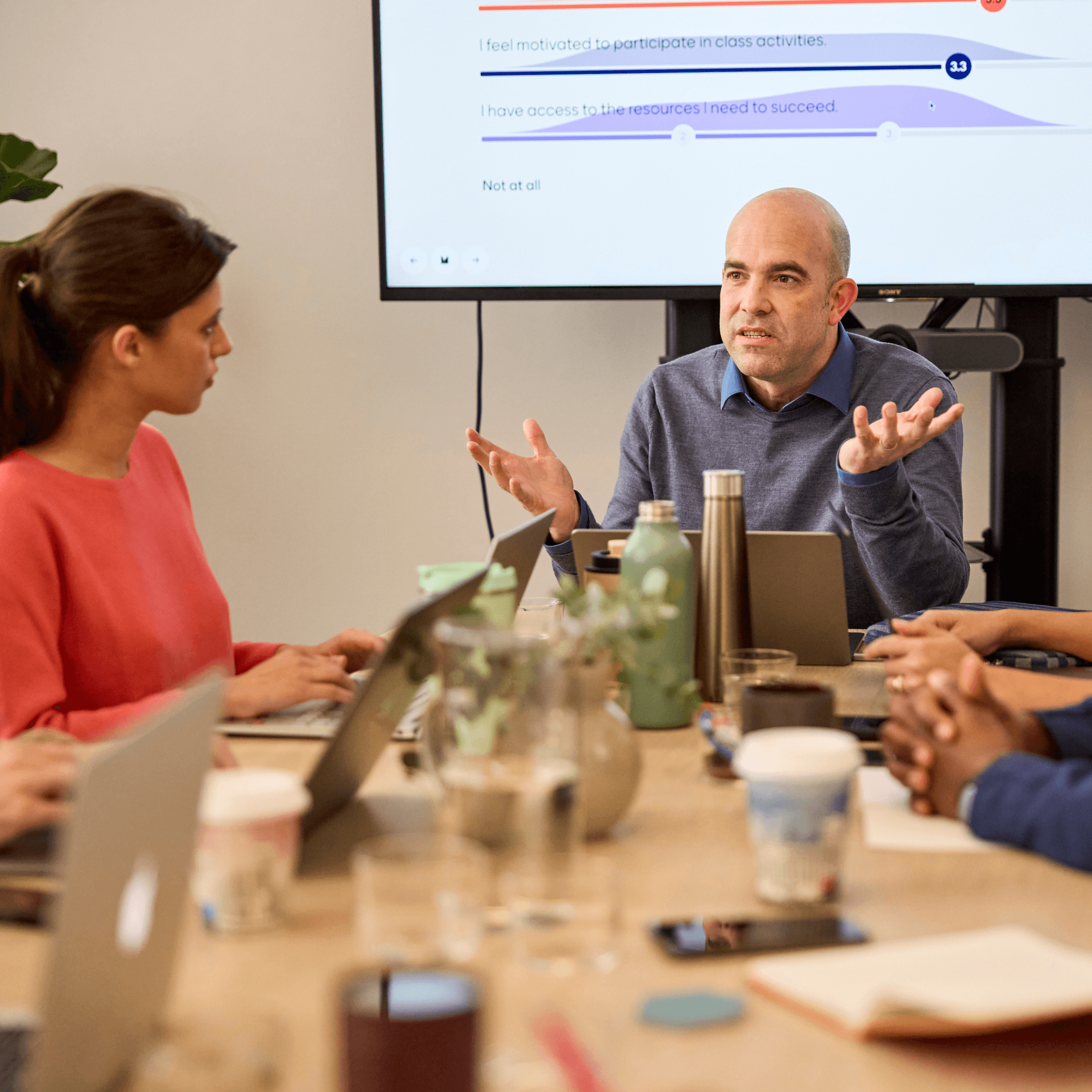 Man speaking in a meeting room with colleagues at a table and survey results on a screen