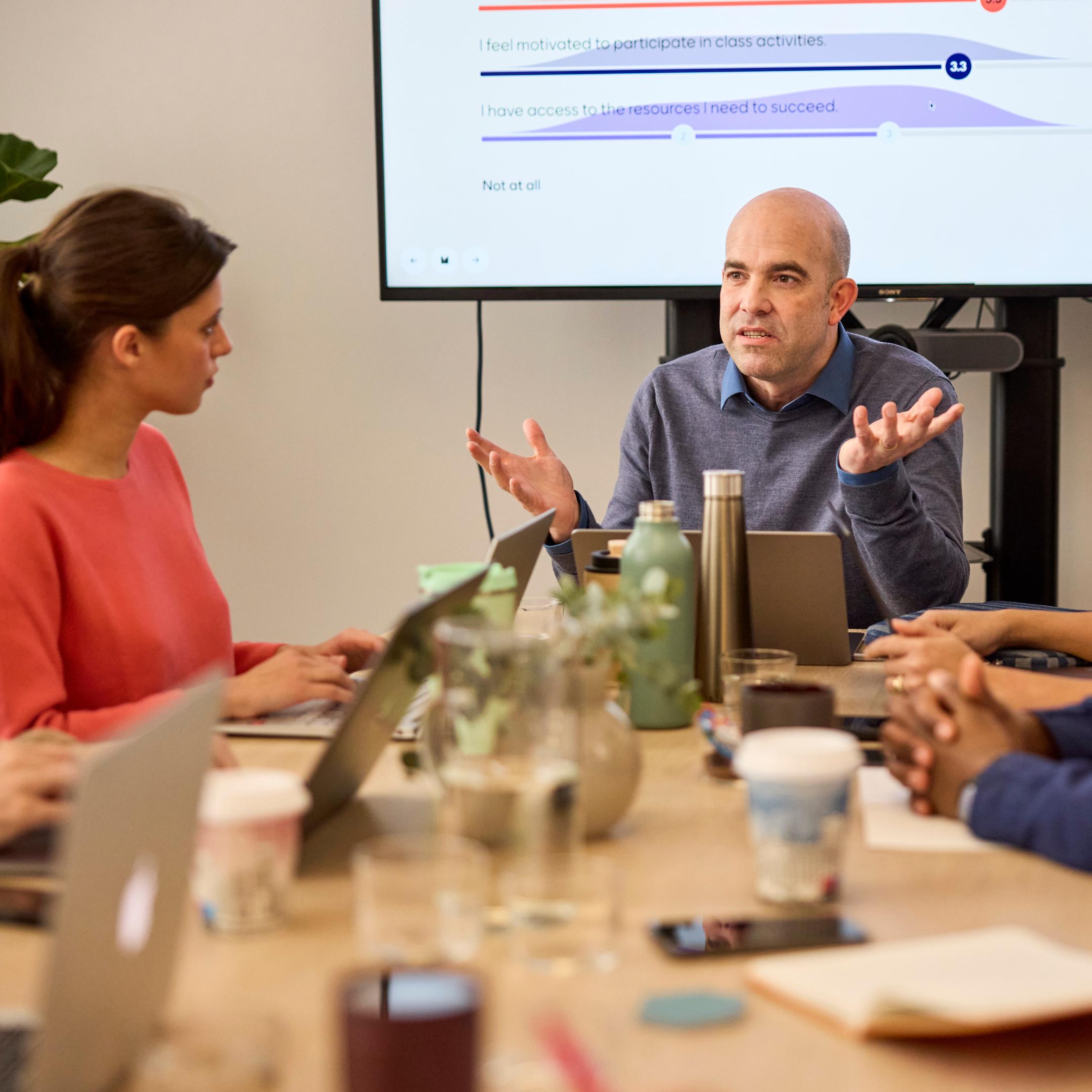 Man speaking in a meeting room with colleagues at a table and survey results on a screen